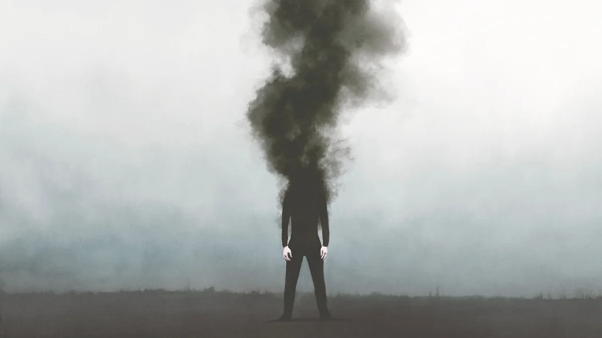 A man in dark clothes standing on a gray background with his head smoking